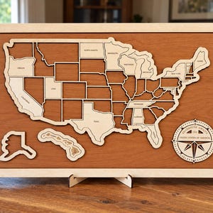May include: A wooden wall map of the United States featuring individual state cutouts mounted on a reddish-brown board with a light wood frame. A circular compass rose emblem in the bottom right corner displays the text Adventure Awaits The United States of America.