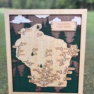 May include: A layered wooden map of Wisconsin State Parks featuring a light wood state outline with engraved park names and tree icons. The design includes dark green pine trees, white clouds, and a brown background, all encased in a light wood frame.