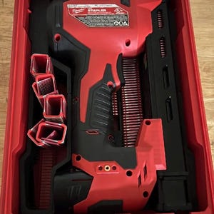 May include: A red and black Milwaukee stapler in a red carrying case. The stapler has a black handle and a red body with the word "STAPLER" printed on it. Several staples are visible in the case.