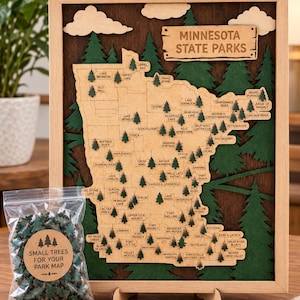 May include: A framed wooden map of Minnesota State Parks featuring layered green trees, a dark wood background, and a clear plastic bag containing small wooden tree markers with a label that reads Small Trees For Your Park Map.