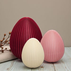 Può includere: Tre oggetti decorativi a forma di uovo di varie dimensioni e colori. Il più grande è rosso scuro, quello medio è rosa tenue e il più piccolo è bianco sporco. Tutti hanno una texture a coste, che suggerisce un design moderno.