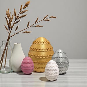 Op de afbeelding: Een stilleven met decoratieve paaseieren in goud, zilver, roze en wit. De eieren hebben een textuur, patroonontwerp. Een takje gedroogde tarwe en twee vazen staan ook in de afbeelding.