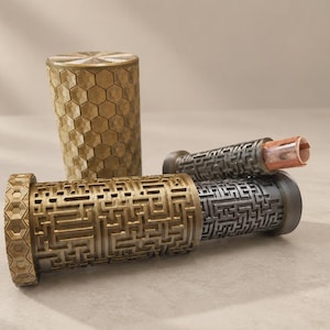 May include: A cylindrical gold-colored container with a hexagonal pattern and a matching lid, positioned next to a horizontal gold and dark gray cylindrical case featuring an intricate maze-like design with a small rolled paper scroll inside.