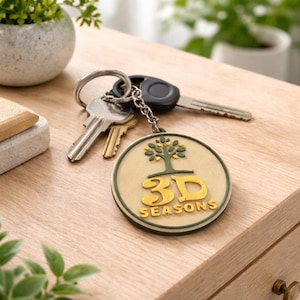 Könnte beinhalten: Ein Schlüsselanhänger mit Schlüsseln auf einer Holzoberfläche. Der Schlüsselanhänger hat einen runden, beigen Anhänger mit grünem Rand, einem Baumdesign und den Worten "3D SEASONS" in Gold. Die Schlüssel sind silber und schwarz.