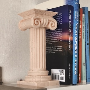 May include: A light peach-coloured Ionic column bookend with detailed carvings. The bookend is placed on a white shelf with several books in the background. The books have titles in English and Hebrew.