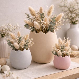 Könnte beinhalten: Drei eiförmige Vasen in Beige, Grau und Rosa, gefüllt mit Trockenblumen und Grünzeug. Die Vasen stehen auf einer neutralen Oberfläche, im Hintergrund weitere Vasen und Deko-Eier.