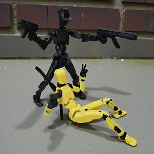 Könnte beinhalten: Zwei bewegliche Actionfiguren. Eine schwarze Figur steht und hält zwei schwarze Spielzeugpistolen. Eine gelbe Figur liegt am Boden, hält ebenfalls eine schwarze Spielzeugpistole und zeigt das Friedenszeichen.