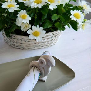 Può includere: Un anello per tovaglioli in ceramica bianca a forma di coniglio che tiene un tovagliolo con un motivo floreale. L'anello per tovaglioli è su un piatto verde. Un cesto di vimini bianco pieno di margherite bianche è sullo sfondo.