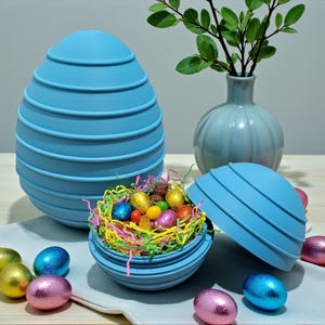 Può includere: Un contenitore a forma di uovo blu chiaro con un design a coste, aperto per rivelare un nido di caramelle pasquali colorate. Diverse uova avvolte in carta stagnola di vari colori sono sparse intorno al contenitore, con un vaso e del verde sullo sfondo.
