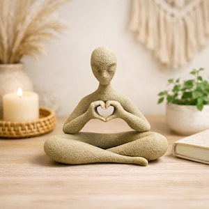 Könnte beinhalten: Beigefarbene Figur einer Person in einer sitzenden Yoga-Pose, wobei die Hände ein Herz formen. Die Figur hat eine strukturierte Oberfläche und steht auf einem Holztisch. Der Hintergrund umfasst eine Kerze, eine Pflanze und ein Buch.