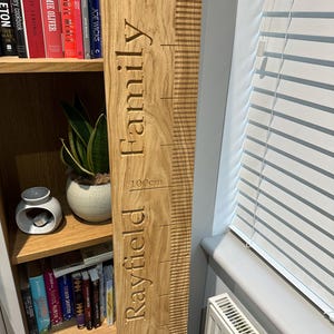 Op de afbeelding: Een houten groeimeter met de tekst "Rayfield Family" en metingen in centimeters. De meter is lichtbruin en heeft een natuurlijke houtnerf.
