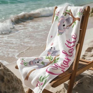Könnte beinhalten: Weißes Strandtuch mit Koala-Bär-Illustrationen auf Ästen, rosa Blumen und dem Namen "Charlotte" in rosa Schrift. Das Handtuch liegt auf einem Holzstrandstuhl an einem Sandstrand.