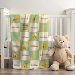 May include: A baby blanket with a bee theme, featuring a repeating pattern of bees, honeycombs, and the name "William" on a green and cream background. A teddy bear sits next to the cot.