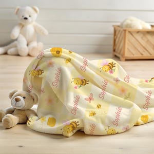 May include: A soft, yellow and white baby blanket with a bee and flower pattern. The blanket has the name "Sophie" printed in pink. A teddy bear and a white bear are in the background.