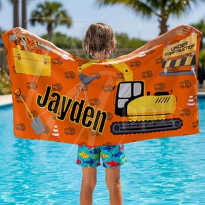 May include: Orange beach towel with a yellow excavator, toolbox, and construction graphics. The towel has the name "Jayden" in large black letters. The words "Under Construction" are also visible. The towel is held up by a person in blue swim shorts.