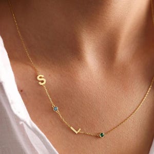 Puede incluir: Un delicado collar de oro con las letras "S" y "L" y tres pequeñas piedras preciosas. Las piedras preciosas son azules y verdes. El collar se lleva sobre una camisa blanca.