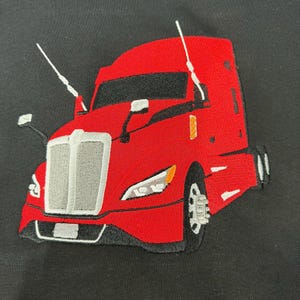 May include: A black t-shirt featuring a detailed red semi-truck embroidery. The truck design includes a prominent silver grille, white antennas, black windows, and visible wheels, centered on the dark fabric background.
