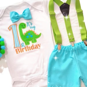 May include: A baby boy's first birthday outfit with a white bodysuit, blue bow tie, green dinosaur graphic, and the text "It's my 1st Birthday". The outfit also includes green suspenders, a blue bow tie, and blue shorts.