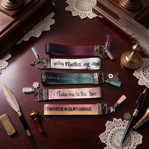May include: Five fabric keychains with phrases like "Flawless, my dear" and "Take me to the ton." Each keychain has a metal clasp and a decorative tassel. The keychains are in shades of purple, white, green, pink, and orange.