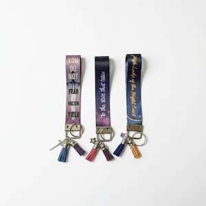 May include: Three keychains with fabric straps and metal hardware. Each strap has a different design and text: "YOU DO NOT FEAR FALTER YIELD", "To the stars that listen", and "High Lady of the Night Court". Each keychain has a tassel.