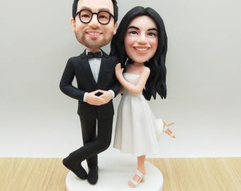 May include: A wedding cake topper featuring a smiling couple. The groom wears a black tuxedo with a bow tie and glasses. The bride is in a white strapless dress. The base is white and has the text "Family Peña Mistry".