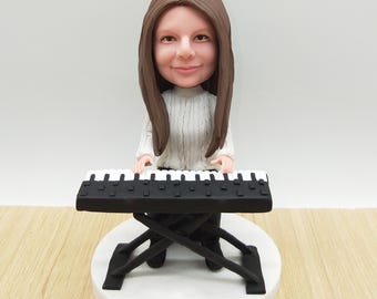 Peut inclure: Figurine d'une personne aux longs cheveux bruns portant un pull blanc, jouant d'un clavier noir et blanc. Le clavier est sur un support noir, et la figurine est sur une base circulaire blanche.