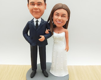 May include: A wedding cake topper featuring a smiling couple. The groom wears a navy suit with a black tie, and the bride is in a white strapless gown with small, raised details. The figures stand on a gray circular base.