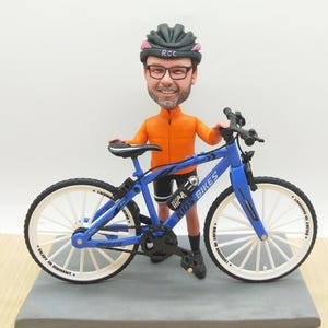 Könnte beinhalten: Ein blaues und weißes Miniaturfahrrad mit dem Text "KIDS BIKES" auf dem Rahmen. Eine Figur eines Mannes in Radsportkleidung, einschließlich Helm und Brille, hält das Fahrrad. Die Räder tragen den Text "ENJOY IN MOMENT".