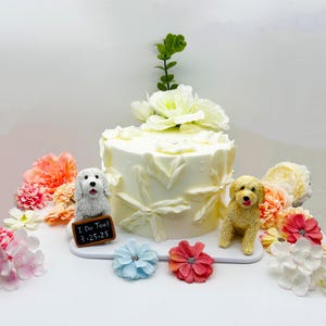 May include: A white wedding cake decorated with white flowers and leaves. Two small dog figurines are on either side of the cake, one white and one yellow. The white dog holds a small chalkboard sign that says "I Do Too! 3.25.25".