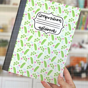 Może przedstawiać: Notatnik kompozycyjny z jasnozieloną okładką z wzorem zielonych ogórków, groszku, różowych kokardek i niebieskich gwiazdek. Zeszyt ma czarny grzbiet i napis "Composition notebook" w ozdobnej ramce.