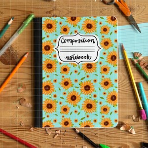 May include: A composition notebook with a sunflower pattern on a light blue background. The notebook has the words "Composition notebook" in a white frame. Pencils, scissors, and glue are scattered around the notebook on a wooden surface.