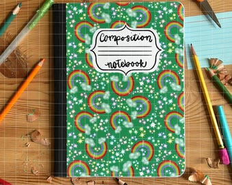 May include: A composition notebook with a green cover featuring a pattern of rainbows, clouds, and stars. The notebook has a black spine and the words "Composition notebook" in a white label. Pencils, scissors, and glue are scattered around the notebook.