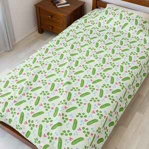 May include: A light green bedspread featuring a repeating pattern of green pickles, pink bows, and blue star-shaped flowers. The bedspread is on a wooden bed frame.