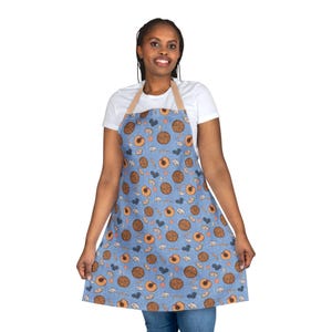 May include: A blue apron featuring a pattern of brown circles, peaches, and small shapes. The apron has a beige neck strap. The apron is being worn.