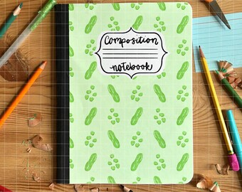 May include: A composition notebook with a light green cover patterned with green pickles and pickle slices. The notebook has the words "Composition notebook" in a decorative label. Pencils, scissors, and glue are scattered around the notebook.