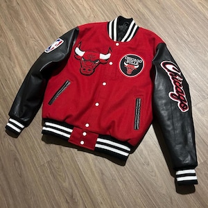 May include: Red and black Chicago Bulls varsity jacket with black leather sleeves. Features the team logo on the chest and a Chicago script on the sleeve. Includes white and black striped cuffs and collar.