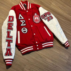 May include: A red and white varsity jacket with white sleeves. The left sleeve has the word "DELTA" in red block letters. The right sleeve has "SPR '23". The jacket has a patch with Greek letters and a crest.