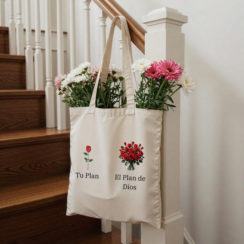 May include: A cream-coloured tote bag filled with white and pink flowers. The bag features two illustrations: a single red rose and a bouquet of red roses. Text on the bag reads "Tu Plan" and "El Plan de Dios". The bag is hanging on a white banister.