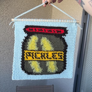 Op de afbeelding: Een blauwe gehaakte wandhanger met een gepixelde afbeelding van een pot met augurken. De pot is groen met een rood deksel en het woord "PICKLES" staat in gele letters.