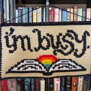 Op de afbeelding: Een gehaakte wandhanger met de tekst "I'm busy" in zwart garen op een beige achtergrond. Een regenboog en een boek zijn onder de tekst gehaakt.