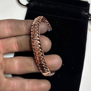 May include: A copper-colored bracelet with a textured, woven design. The bracelet is held in a hand, and a black drawstring bag is in the background. The bracelet's design is made of repeating, curved segments.
