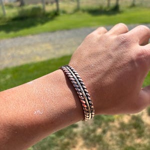 Peut inclure: Un bracelet manchette composé d'une bande centrale lisse encadrée par deux bandes torsadées en forme de corde. Le bracelet est fabriqué en métal avec des finitions alternées de couleur cuivre et laiton, créant un aspect superposé.