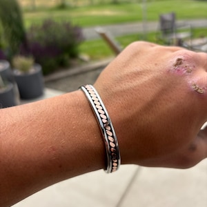 May include: A cuff bracelet featuring a polished silver-tone outer band and a central inlay of copper-colored oval links. The metal bracelet has a smooth, rounded profile and a continuous pattern of repeating metallic shapes along its entire length.