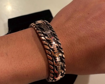 May include: A silver-toned bracelet with a woven design is displayed on a wrist. The bracelet has a thick, textured appearance with intricate detailing. The background includes a black pouch and a light-colored surface.