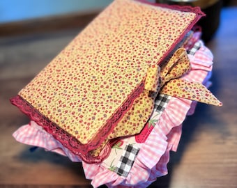 May include: A fabric-covered book featuring a yellow floral print with a dark red lace trim and a matching fabric bow. The underlying layers include a pink and white gingham ruffle, a black and white buffalo check pattern, and a strawberry print.