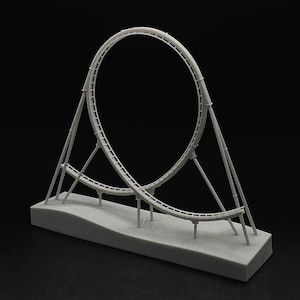 Peut inclure: Maquette grise d'un modèle de montagnes russes avec une conception en boucle. Le modèle est posé sur une base rectangulaire et porte le texte "THRILL MAKERS" en bas. Les supports et la piste du coaster sont visibles sur fond noir.
