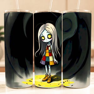 May include: A set of three stainless steel tumblers with a spooky cartoon girl design. The girl has pale skin, long white hair, and a colorful patchwork dress. The background is black with a swirling design, and the base is yellow.