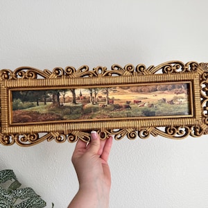 May include: A rectangular landscape painting featuring a pastoral scene with grazing cattle in a field, enclosed within an ornate, decorative gold-colored frame with intricate scrollwork and textured detailing along the edges.
