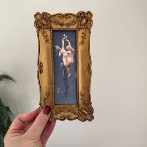 May include: A rectangular gold-toned frame with ornate, raised floral and scrollwork detailing. The frame encloses a vertical print depicting two figures floating against a dark blue background, with one figure reaching upward toward small, glowing white stars.