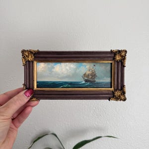以下が含まれることがあります： 曇り空の下、深い青色の海に浮かぶ帆船を描いた長方形の額入り油絵。フレームはダークブラウンで、四隅に装飾的な金色のアクセントがあり、内側には細い金色の縁取りが施されており、クラシックで詳細な外観を作り出しています。